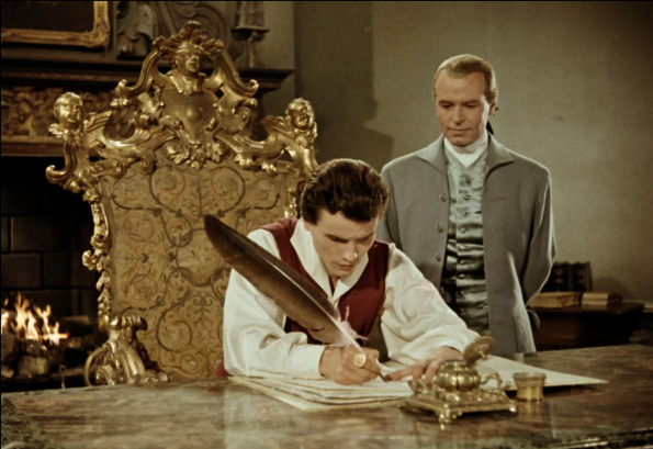 Ausschnitt aus dem Film "Herrscher ohne Krone". Umgeben von prunkvollem Mobiliar sitzt König Christian VII. mit einer Schreibfeder über Papiere gebeugt am Schreibtisch, im Hintergrund steht Struensee und blick ihm über die Schulter.