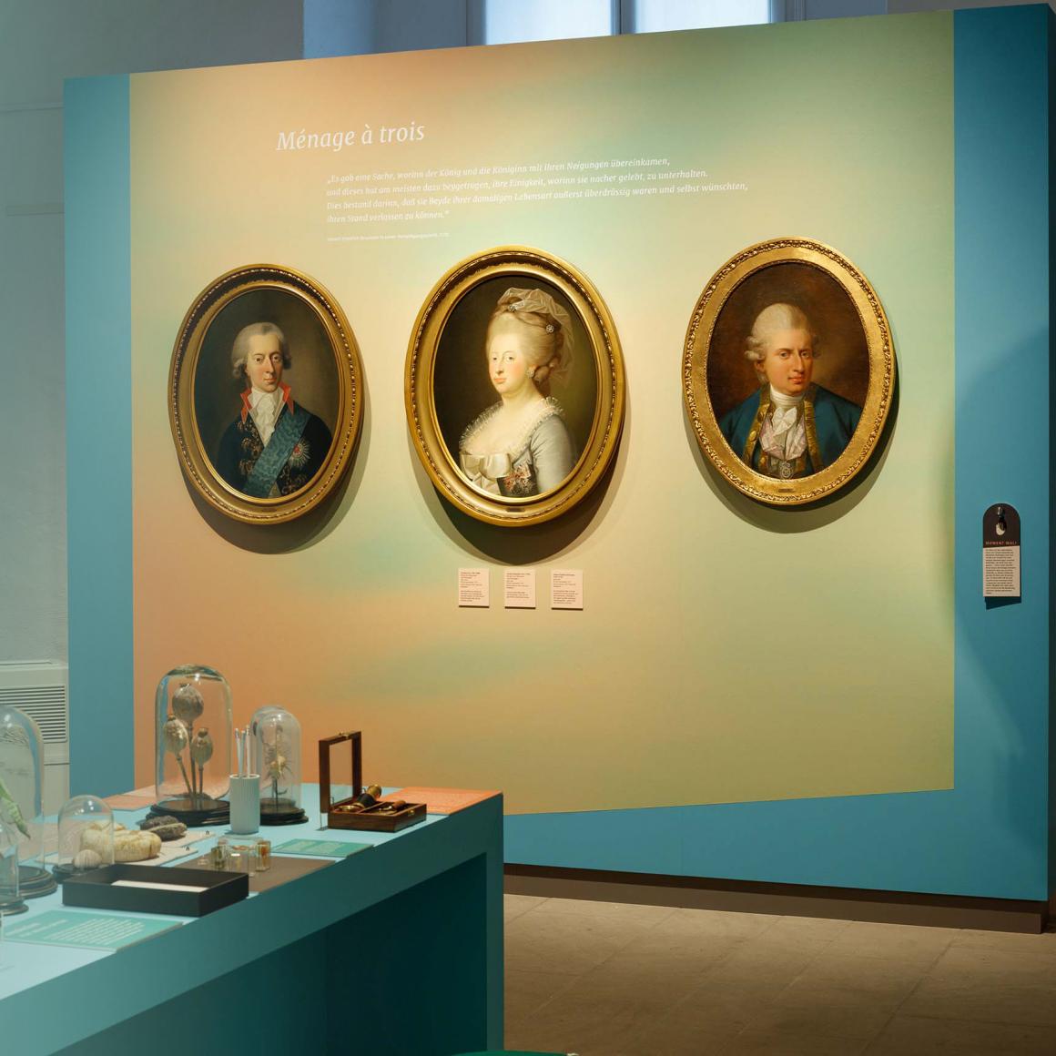 An der Wand drei ovale Porträts von Christian VII., Caroline Mathilde und Johann Friedrich Struensee, links ein Tisch mit kleinen Exponaten